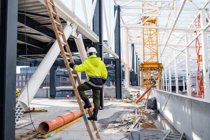 A man in a yellow jacket and hard hat climbs a ladder on a work site.