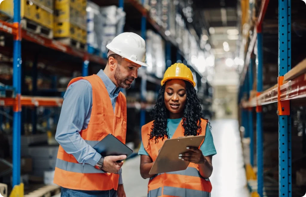 A man and woman in a warehouse consult about something on a clipboard.