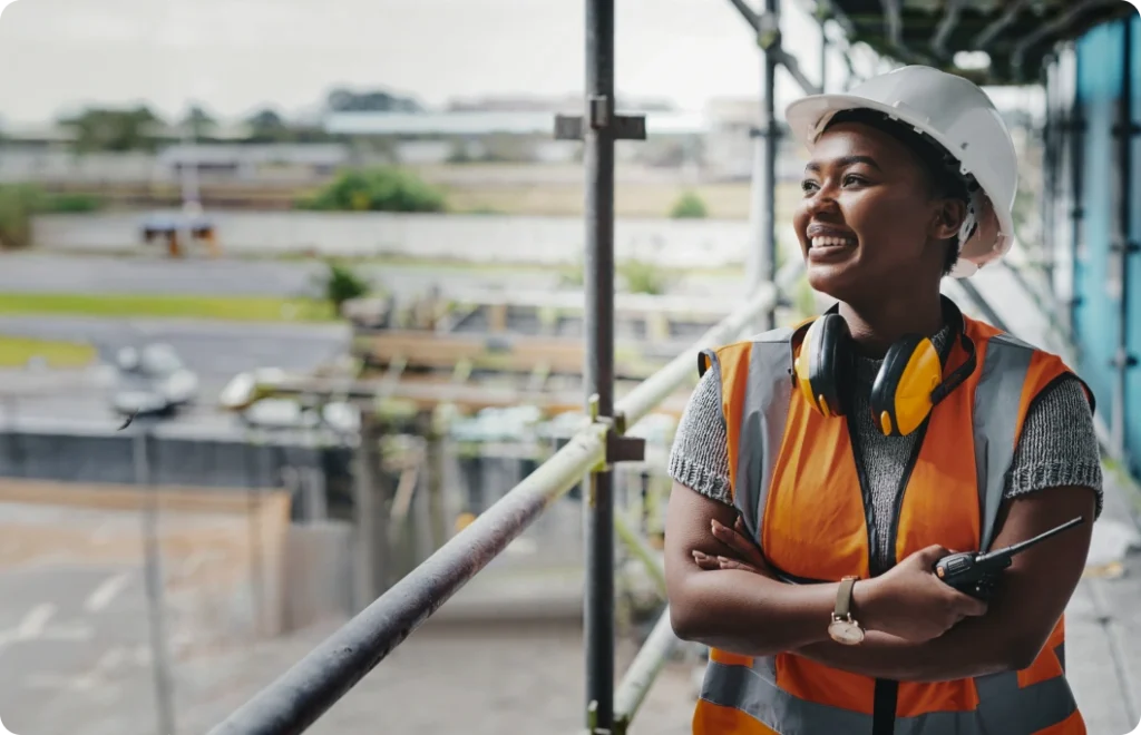 A woman in an orange vest and white hard hat crosses her arms and looks to her right.