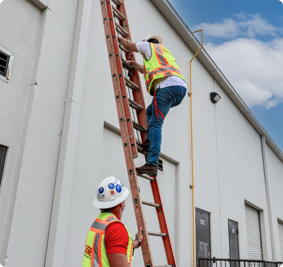 A man in a yellow vest climbs a ladder while another man holds the ladder still below him.